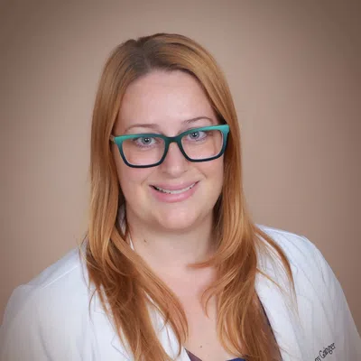 Smiling female doctor with long red hair and teal glasses wearing a white coat against a beige background