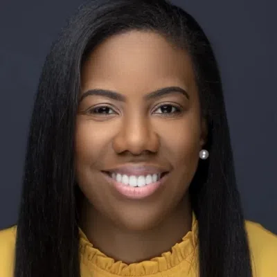 Smiling woman with straight black hair wearing a yellow top and pearl earrings against a dark background