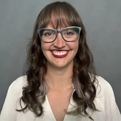 Smiling woman with long curly hair and glasses wearing a white shirt against a gray background