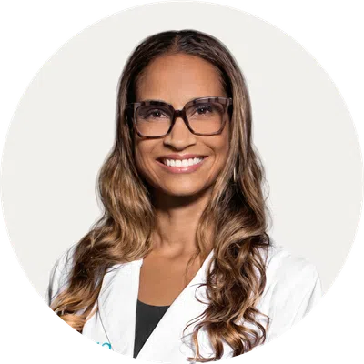Smiling woman with long wavy hair wearing glasses and a white lab coat against a plain background