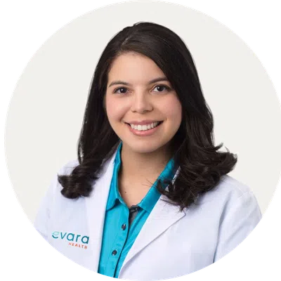 Smiling female healthcare professional wearing white coat and teal shirt with evara health logo on coat.
