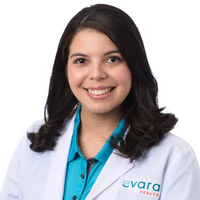 Smiling female healthcare professional wearing lab coat and teal shirt with Evara Health logo on chest.
