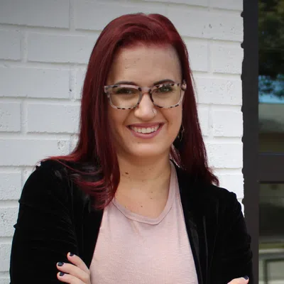 Smiling woman with red hair, glasses, and tattooed arm standing with crossed arms by a white brick wall.