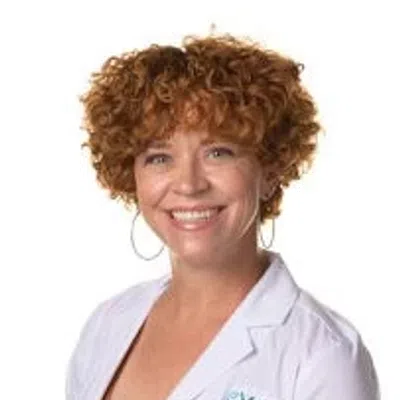 Smiling woman with curly red hair wearing a white Evora medical coat and hoop earrings against white background