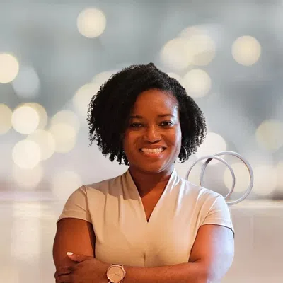 Smiling woman with curly hair and crossed arms wearing a beige top and watch against a blurred light background.