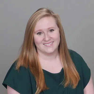 Smiling woman with long blonde hair wearing a dark green top sitting against a plain gray background.
