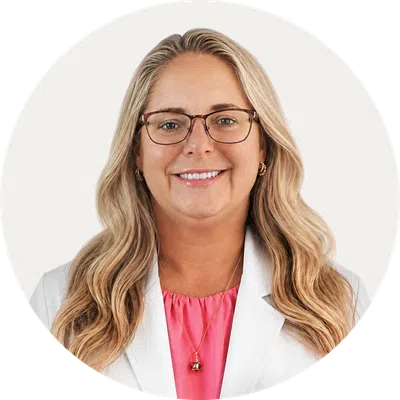 Smiling woman with long blonde hair wearing glasses, white blazer, and pink top in professional headshot.