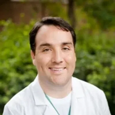 Smiling male doctor wearing a white lab coat with a blurred green outdoor background.