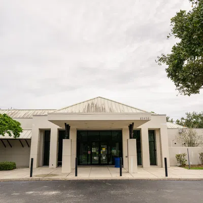 Modern white building with a peaked roof, glass entrance, and surrounding trees under cloudy sky
