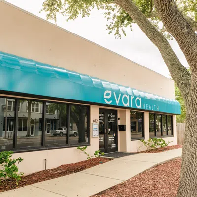 Exterior of Evara Health clinic with blue awning, large windows, and surrounding greenery on sidewalk.