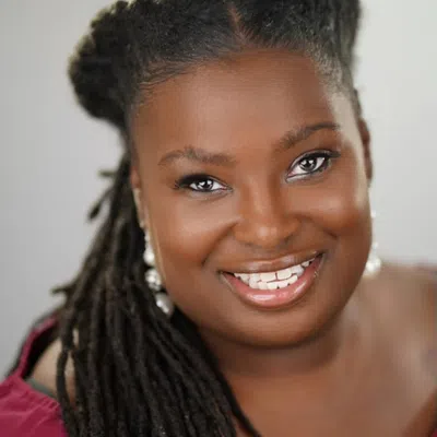 Close-up portrait of a smiling Black woman with long dreadlocks wearing pearl earrings and a burgundy top.