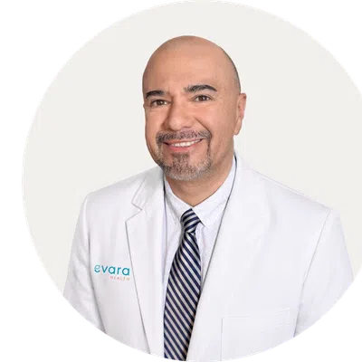 Smiling bald man with goatee wearing a navy blazer, blue shirt, and red-striped tie against a beige background.