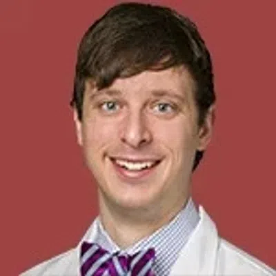 Young man smiling wearing a white lab coat, striped shirt, and purple bow tie with a red background
