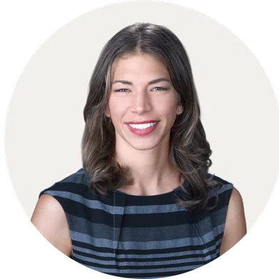 Smiling woman with wavy brown hair wearing a black and gray striped sleeveless top on a white background.
