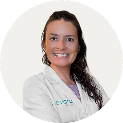 Smiling female healthcare professional with dark curly hair wearing a white Evara Health lab coat.