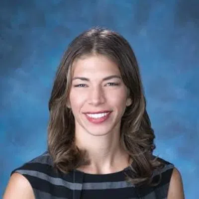 Smiling woman with shoulder-length brown hair wearing a black and gray striped sleeveless top against blue background
