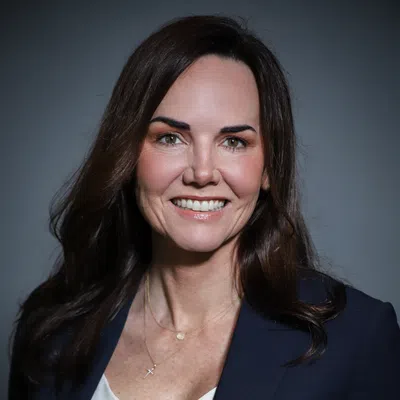 Smiling woman with long brown hair wearing a navy blazer and white top against a gray background.
