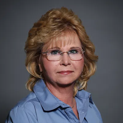 Portrait of a middle-aged woman with blonde hair, glasses, and a blue collared shirt against a gray background.