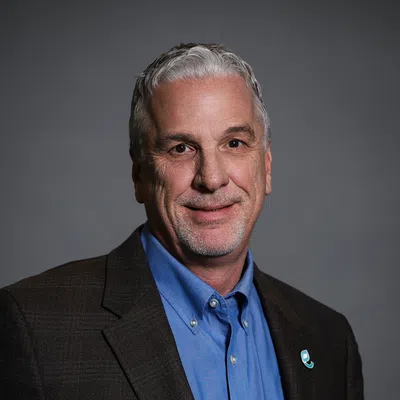 Smiling mature man with gray hair in blue shirt and dark blazer against a plain gray background.