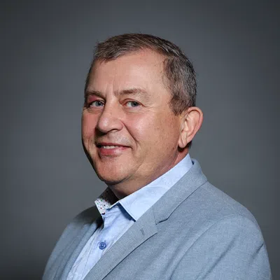 Middle-aged man in light blue blazer and patterned shirt smiling against dark gray background