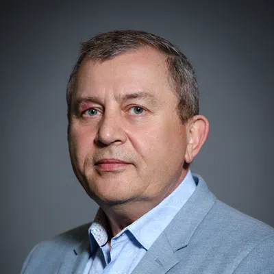 Middle-aged man in a light blue patterned shirt and gray blazer against a dark gray background.