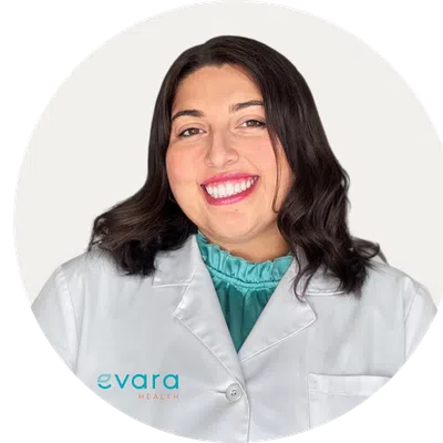Smiling woman in white medical coat with teal blouse, Evara Health logo visible on coat pocket.