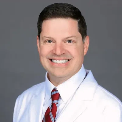 Smiling male nurse practitioner wearing white coat and red striped tie against gray background