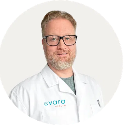 Smiling man with light red hair and beard wearing a white lab coat with Evara Health logo