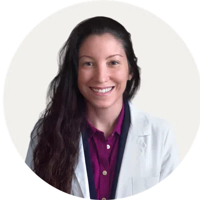 Smiling woman with long dark hair wearing a white lab coat and purple shirt on a white background