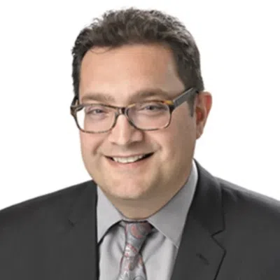 Smiling man wearing glasses, a gray shirt, paisley patterned tie, and dark suit jacket on white background