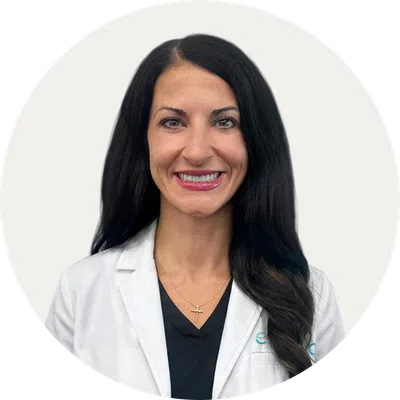 Smiling woman with long black hair wearing a white lab coat and black shirt against a plain background