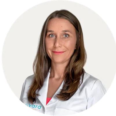 Smiling woman with long brown hair wearing a white lab coat against a plain white background.