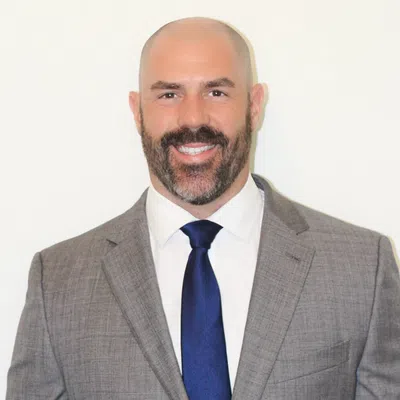 Smiling bald man with beard wearing gray suit, white shirt, and blue tie against white background
