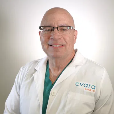 Smiling bald male doctor wearing glasses and Evara Health white coat over teal scrubs against a plain background.