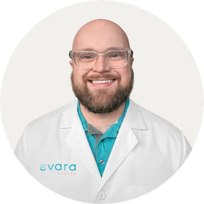 Smiling bald man with glasses wearing a white Evara Health lab coat and coral shirt on a white background
