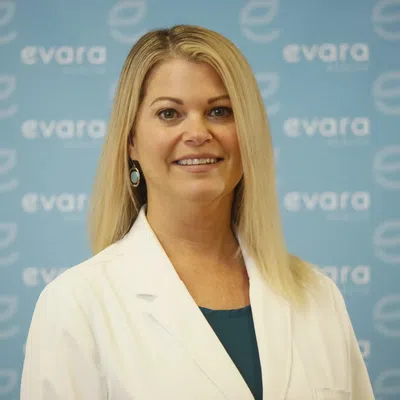 Smiling woman in white medical coat standing in front of blue Evara Health logo backdrop.