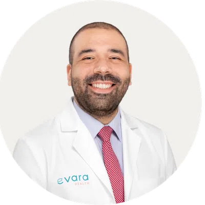 Smiling male doctor wearing white coat and red tie with Evara Health logo on coat against white background