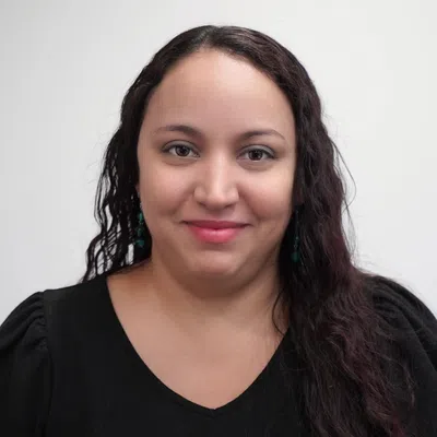 Smiling woman with long curly dark hair wearing black top and turquoise earrings, neutral gray background.
