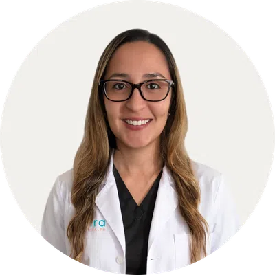 Smiling female doctor with long hair and glasses wearing a white lab coat over black scrubs on white background