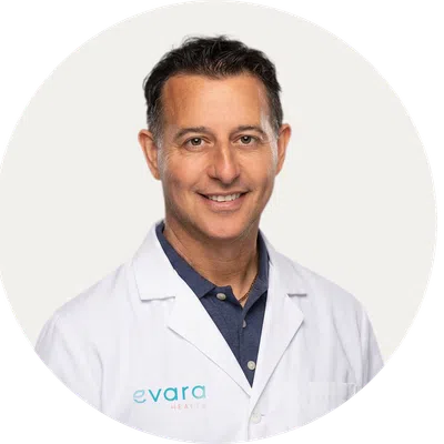 Smiling male doctor wearing a white lab coat with Evard Health logo on a plain light background.