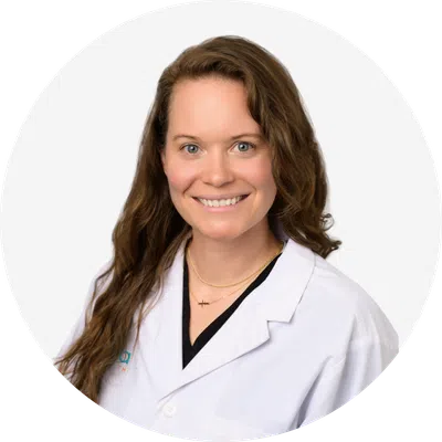 Smiling female doctor with long brown hair wearing a white lab coat against a white background.