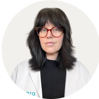 Woman with dark layered hair and red glasses wearing a white coat over black top in a circular frame.