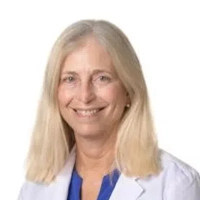 Smiling female doctor with blonde hair wearing a white lab coat and blue scrubs against a white background