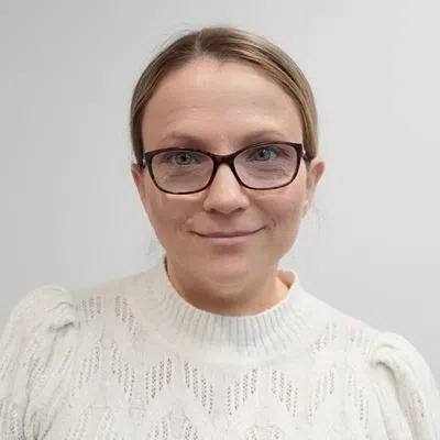 Smiling woman wearing glasses and a white knitted sweater against a plain light background.