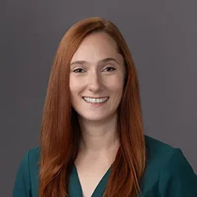 Smiling woman with long red hair wearing a dark green zip-up top against a gray background