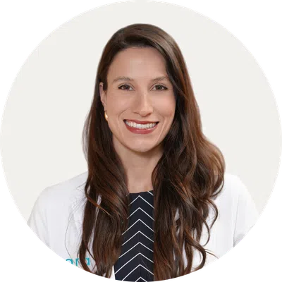 Smiling woman with long brown hair wearing a white coat and striped black shirt against a white background