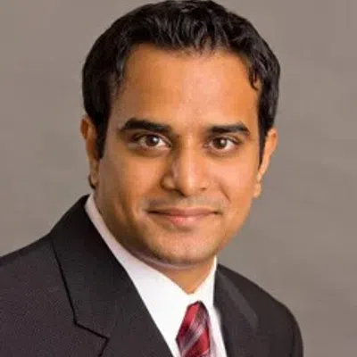 Professional headshot of a man in a dark suit with a red striped tie on a gray background.