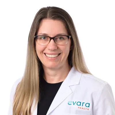 Smiling woman with long blonde hair wearing glasses and a white Evara Health lab coat on a white background