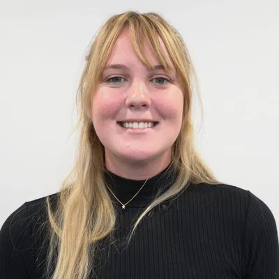 Smiling young woman with long blonde hair wearing a black turtleneck and star necklace against a plain background