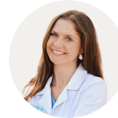 Smiling female doctor with long brown hair wearing a white lab coat against a plain background
